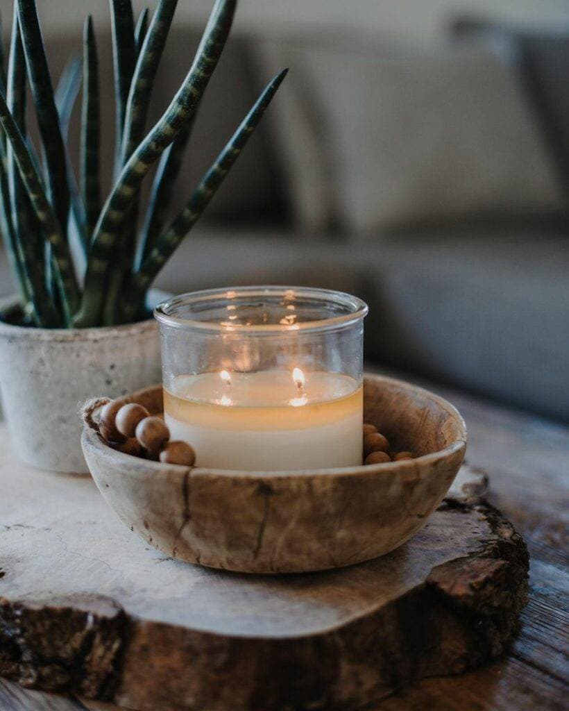 A lit Old Line Candle Co. candle in a wooden bowl with wooden beads, beside a snake plant on a rustic wood slab.