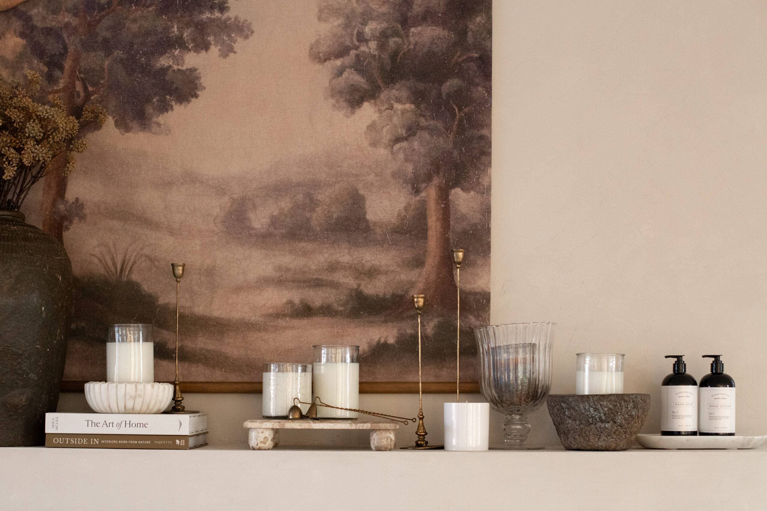 A styled mantel with lit Old Line Candle Co. candles, brass candlesticks, dried flowers, and hand soap and lotion bottles, set against a muted landscape mural.